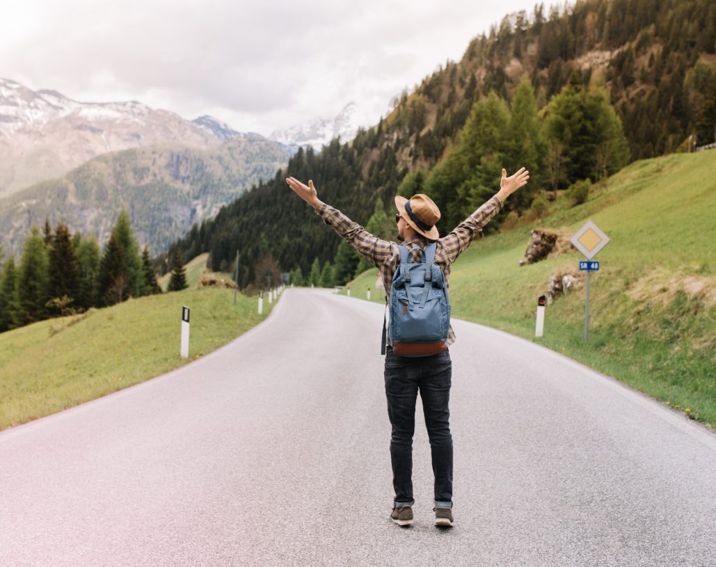 Full-length portrait of blissful man with backpack emotionally posing with hands up and hitchhiking on the highway. Alone traveler in trendy hat going to distant Alps enjoying mountain landscape.